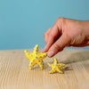 A hand holds a small starfish from the Bumbu Handmade Wooden Yellow Starfish Set, featuring yellow wood with blue spots beside a matching larger piece on a light wooden surface and blue background—ideal for Montessori learning.