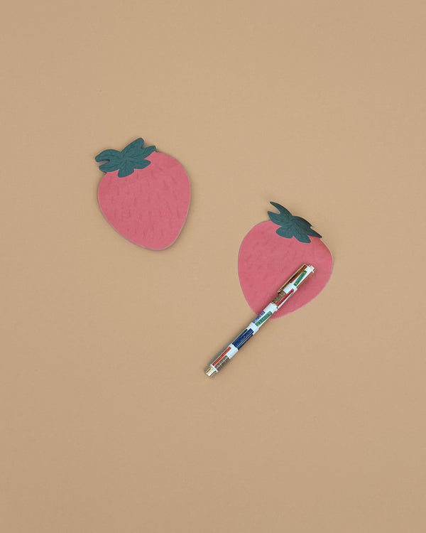 Two Strawberry Sticky Notes, pink with green tops and a fun die-cut strawberry shape, rest on a beige surface. A multicolored pen is placed diagonally across one of the adhesive sheets.