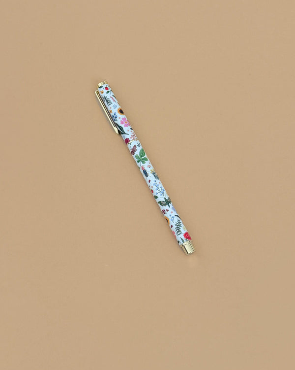 The Curio Pen, a 0.5mm gel ink rollerball with a floral design in red, green, and blue accents, rests diagonally on a plain light brown surface.