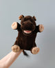 The Hippo Puppet, featuring a plush hand design, is showcased against a simple light grey backdrop. The puppet boasts a rich brown color with lighter beige accents on its feet, mouth, and nostrils. It also has plush ears, a wide mouth with a pink tongue, and small brown eyes that contribute to its charmingly realistic appearance.