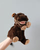 The Hippo Puppet, featuring a plush hand design, is showcased against a simple light grey backdrop. The puppet boasts a rich brown color with lighter beige accents on its feet, mouth, and nostrils. It also has plush ears, a wide mouth with a pink tongue, and small brown eyes that contribute to its charmingly realistic appearance.
