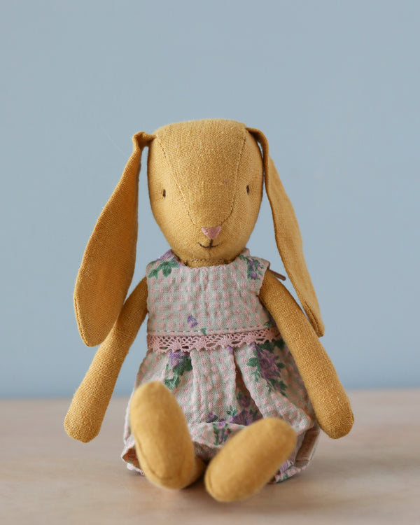 A Maileg Bunny Size 2 in dusty yellow, wearing a dress, sits on a light wooden surface against a plain light blue background.