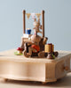 A Wooden Bunny Music Box, crafted from sustainably sourced wood, featuring a small figure in orange taking a photo, a cake with a number one on top, and colorful decorations on a light wooden table.