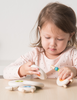 A young girl focuses intently on playing with a Lacing Wooden Sheep on a table, carefully examining one in her hands.