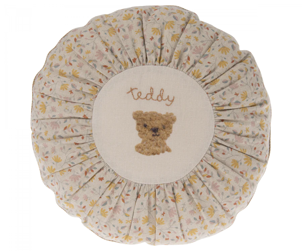 A round, floral-patterned Maileg Cushion, Small with a central embroidery of a teddy bear's face and the word "teddy" stitched above it in gentle script.