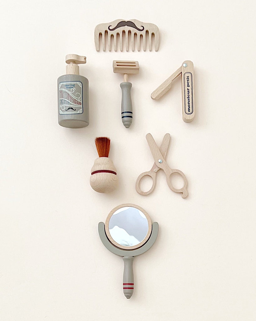 An array of Pretend Play Wooden Barber Set neatly arranged on a light background, including a shaving brush, razor, after-shave bottle, a comb, tweezers, nail clippers, scissors, and a hand