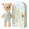 Cuddle + Kind Tiny Charlie the Honey Bear, in pastel green overalls, stands by an arched box adorned with whimsical soft-colored flowers, a bear, and bees. Crafted using fair trade practices and filled with hypoallergenic polyfill, it's ideal for gentle play.
