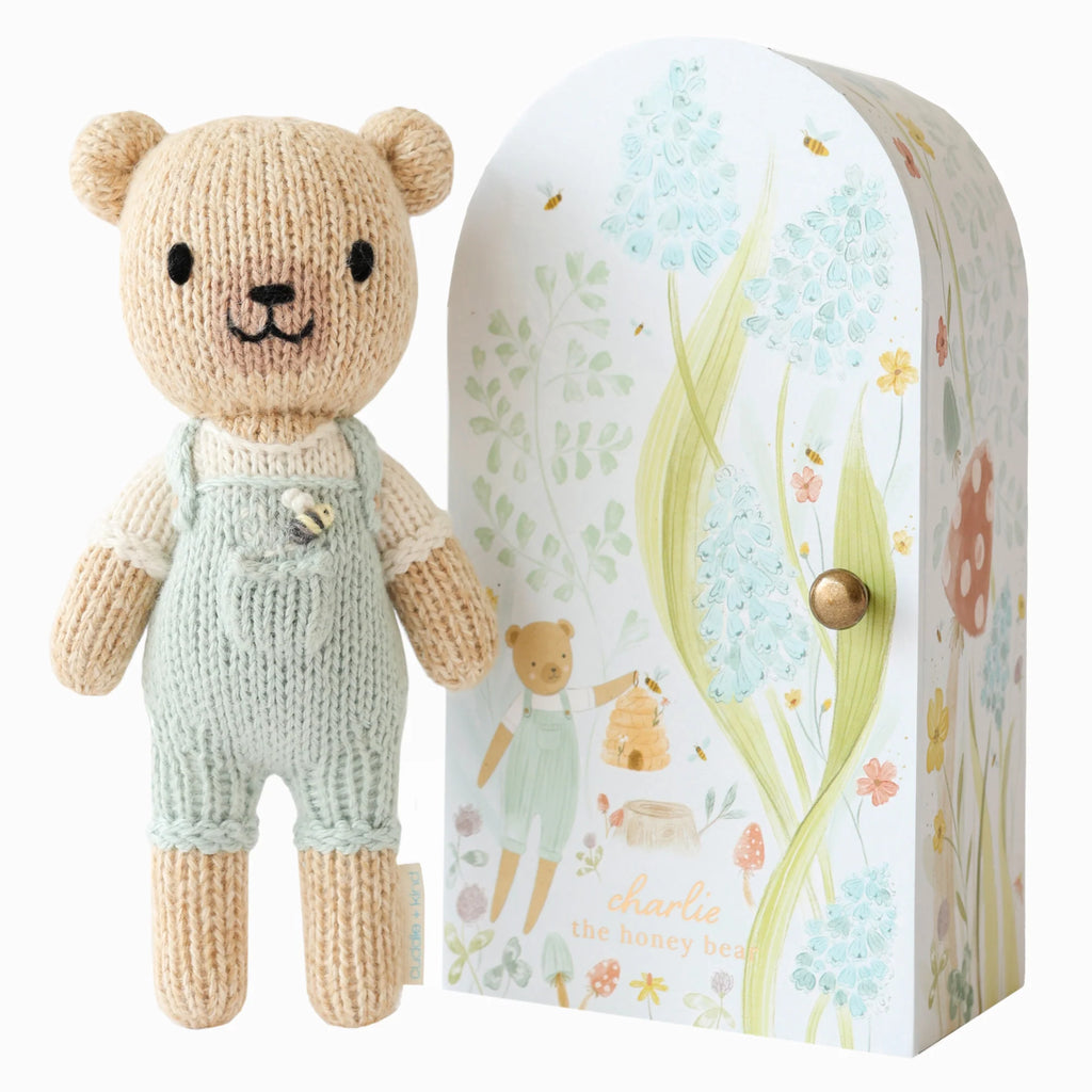 Cuddle + Kind Tiny Charlie the Honey Bear, in pastel green overalls, stands by an arched box adorned with whimsical soft-colored flowers, a bear, and bees. Crafted using fair trade practices and filled with hypoallergenic polyfill, it's ideal for gentle play.