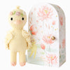 The Cuddle + Kind Tiny Flora the Duckling in mauve, adorned with floral accessories, stands beside a decorative box featuring pastel lotus flowers and duckling illustrations. This charming, sustainable toy supports fair trade practices.