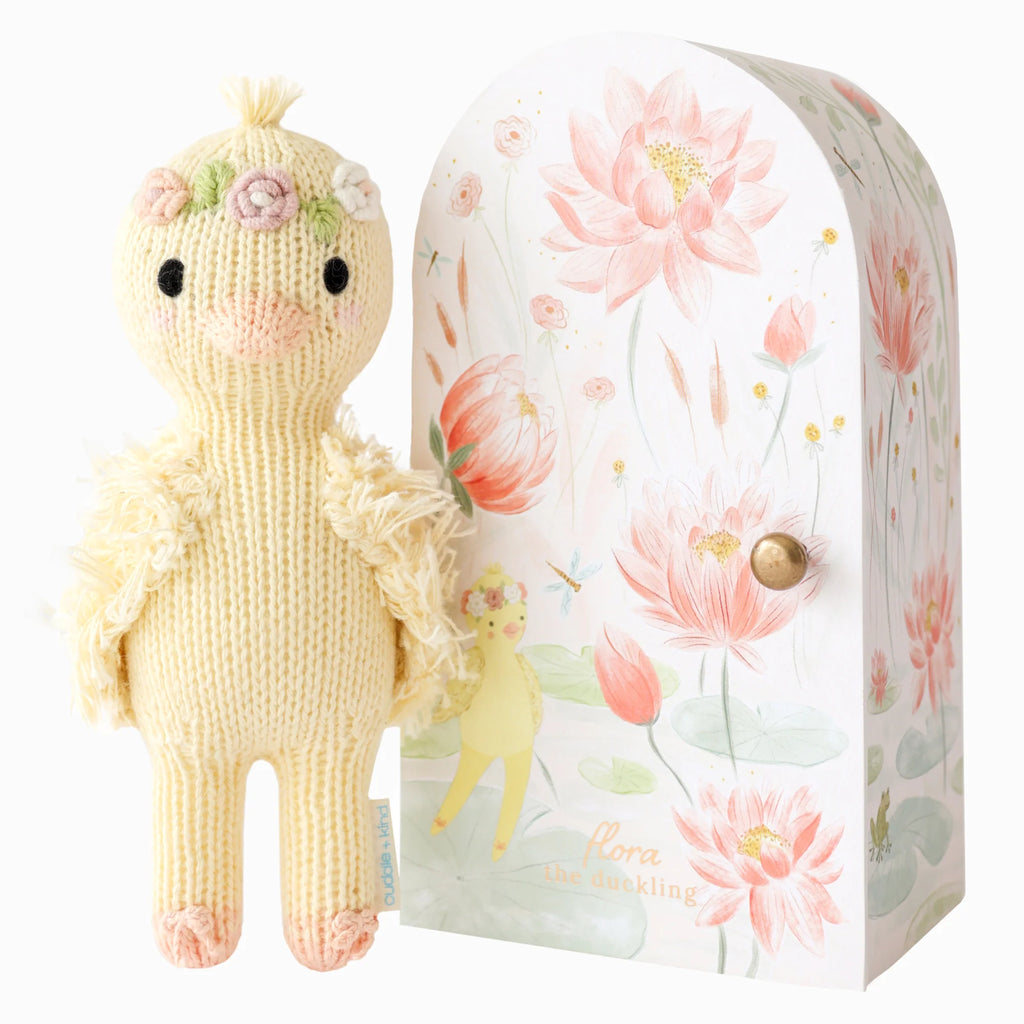 The Cuddle + Kind Tiny Flora the Duckling in mauve, adorned with floral accessories, stands beside a decorative box featuring pastel lotus flowers and duckling illustrations. This charming, sustainable toy supports fair trade practices.