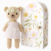 The Cuddle + Kind Tiny Olivia the Honey Bear is a hand-knit toy wearing a flower crown and white dress, standing next to a decorative box with floral designs and an illustration of "Olivia the Honey Bear." Crafted sustainably, the flowers are white, yellow, and pink.