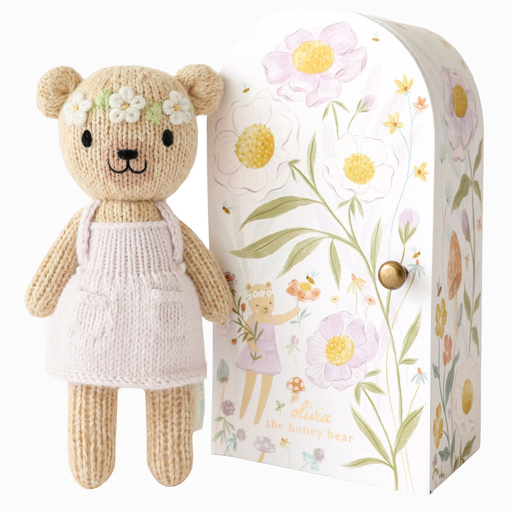 The Cuddle + Kind Tiny Olivia the Honey Bear is a hand-knit toy wearing a flower crown and white dress, standing next to a decorative box with floral designs and an illustration of "Olivia the Honey Bear." Crafted sustainably, the flowers are white, yellow, and pink.