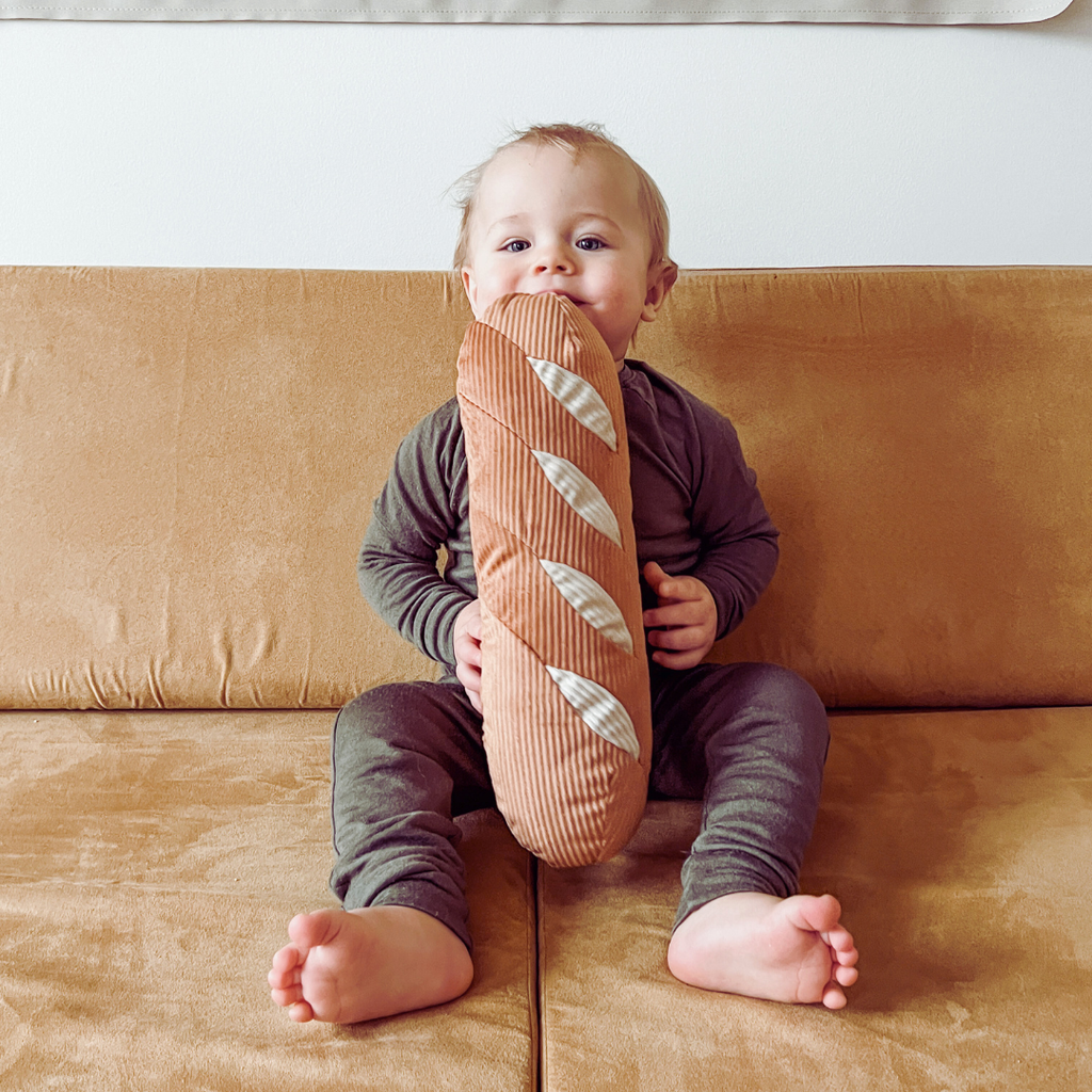 A baby in a dark onesie sits on a tan couch, smiling with delight while holding and playfully biting the Tummy Time Baguette—a large plush pillow ideal for pretend play bakery fun.