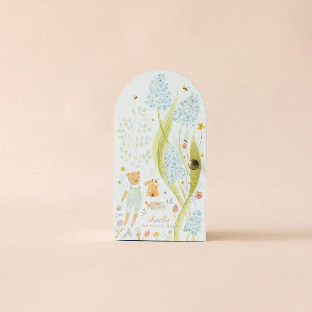 An illustrated card featuring floral designs and a small bear on a neutral background, with the text "Tiny Charlie the Honey Bear." The pastel flowers and bees adorn its curved top with a small knob, showcasing fair trade practices. Product: Cuddle + Kind Tiny Charlie the Honey Bear.