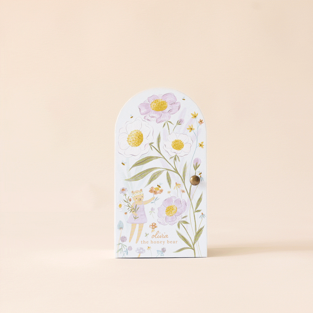 A small decorative door with pastel floral designs stands on a neutral backdrop, opening to reveal the phrase "Cuddle + Kind Tiny Olivia the Honey Bear," evocative of sustainable fair trade.