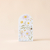 A small decorative door with pastel floral designs stands on a neutral backdrop, opening to reveal the phrase "Cuddle + Kind Tiny Olivia the Honey Bear," evocative of sustainable fair trade.