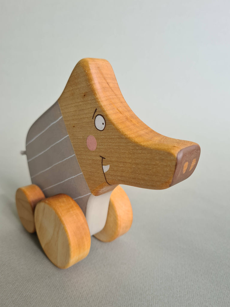 The Pull and Push Toy Little Wild Boar features a smiling face and wheels, painted in natural wood tones with stripes. Standing against a soft green background, this handcrafted wooden toy is made from sustainable materials, bringing joy while being eco-friendly.