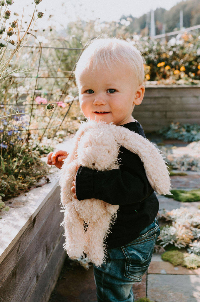 Amidst flowers under a clear sky, a blonde toddler in a black sweater and jeans smiles in the garden while holding Harey the Bunny, a fluffy stuffed toy with embroidered features.
