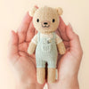 Two hands tenderly cradle Cuddle + Kind's Tiny Charlie the Honey Bear, a fair-trade handmade teddy filled with hypoallergenic polyfill. Dressed in mint overalls and a cream shirt, its embroidered face features black eyes and a tiny smile, all set against a neutral backdrop.