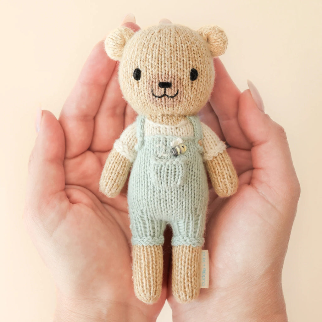 Two hands tenderly cradle Cuddle + Kind's Tiny Charlie the Honey Bear, a fair-trade handmade teddy filled with hypoallergenic polyfill. Dressed in mint overalls and a cream shirt, its embroidered face features black eyes and a tiny smile, all set against a neutral backdrop.