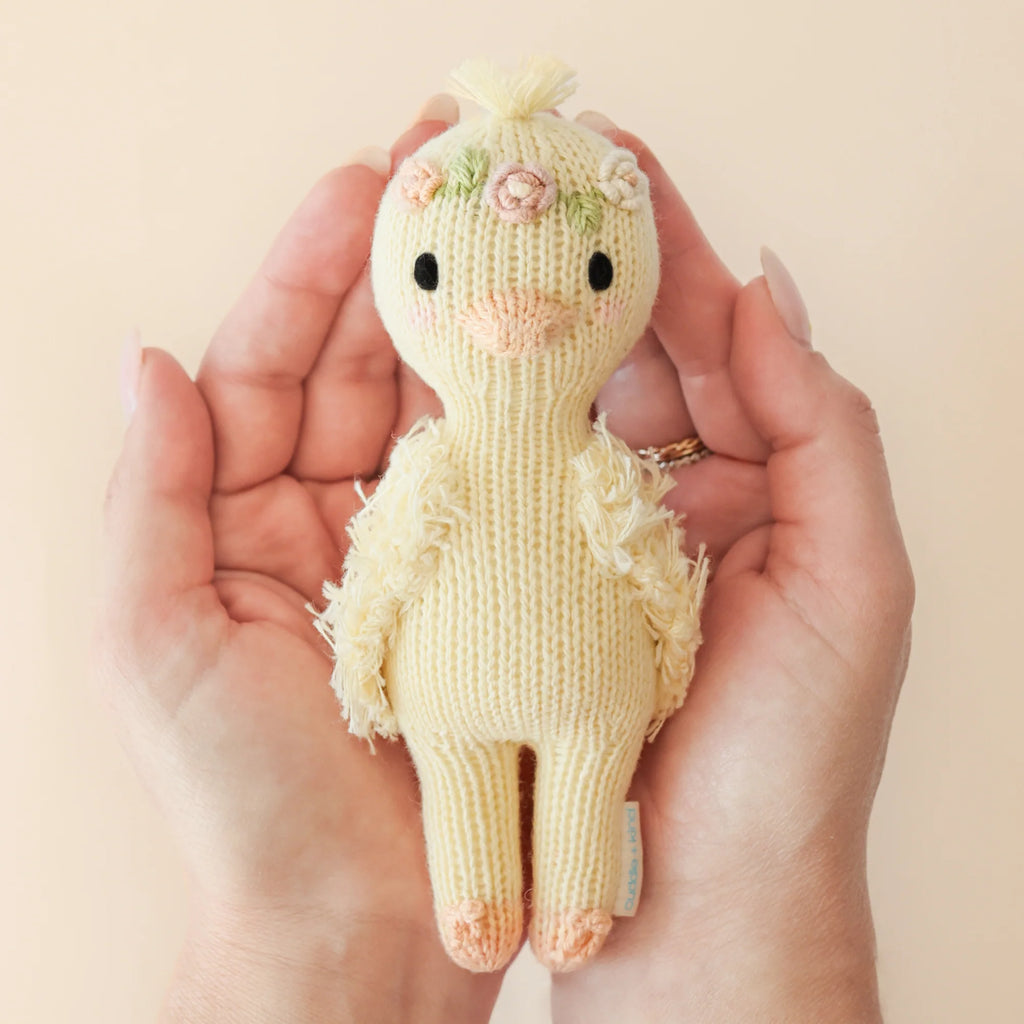 Hands gently cradle Cuddle + Kind's Tiny Flora the Duckling in mauve, crafted using fair trade practices. This knitted bird features floral accents on its head, black eyes, an orange beak, and soft textures against a plain light background.