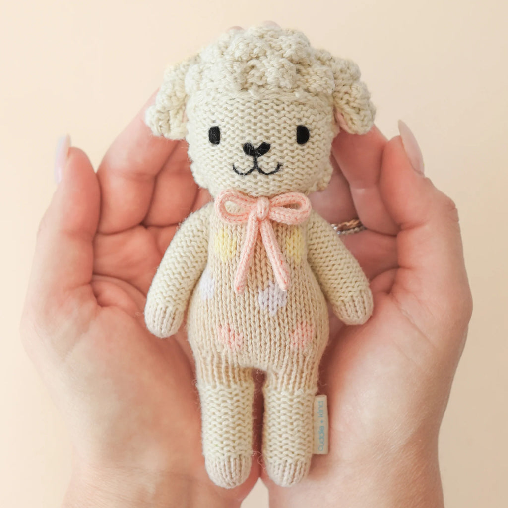 Gently holding the Cuddle + Kind Tiny Lucy the Lamb, a fair trade knitted plushie with black eyes and nose, is a soft cream lamb featuring a pink bow and heart patterns—a charming example of sustainable toys.