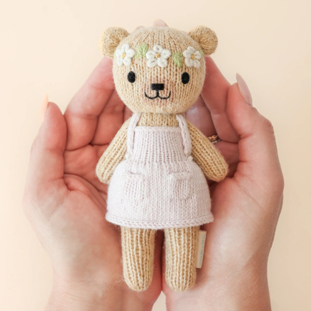 Hands cradle a charming, hypoallergenic Cuddle + Kind Tiny Olivia the Honey Bear. With a beige body, white apron, and flower headband, the bear's warm smile shines against a soft peach background, epitomizing cozy fair trade craftsmanship.