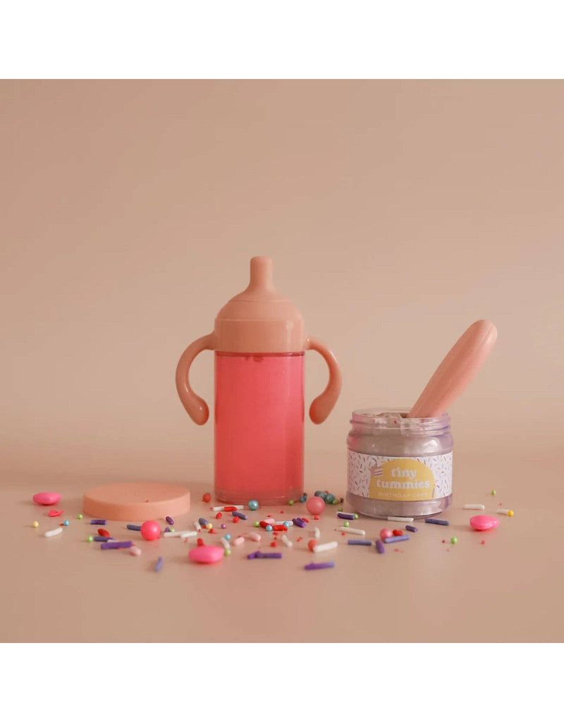 A pink sippy cup with a lid sits on a beige surface amidst colorful sprinkles, resembling a magical strawberry milk scene. Beside it is an open jar labeled "tiny tummies," similar to a birthday cake jar, complete with a spoon inside and the lid off, creating delightful realistic play from the My tiny birthday set - Tiny Harlow.
