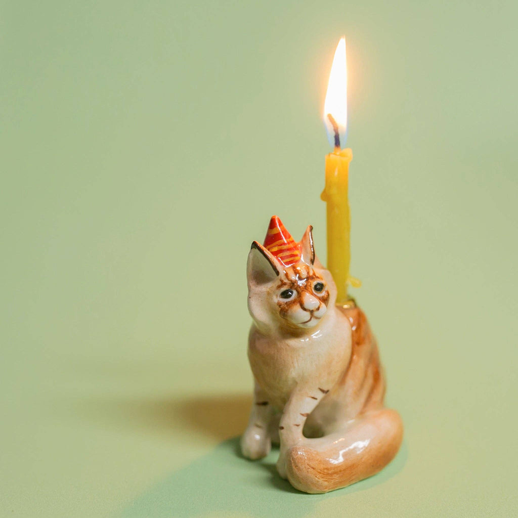 The Maine Coon Cat Cake Topper is a ceramic figurine set against a green background, featuring a cream-colored cat with orange details and a red party hat. A yellow-lit candle on its back adds a softly glowing flame, enhancing the festive ambiance for cat-themed parties.