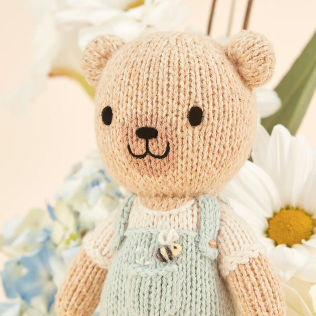 Cuddle + Kind's Tiny Charlie the Honey Bear, wearing light blue overalls with a tiny bee design, stands in front of blurred daisies and light blue flowers. This handmade knitted teddy bear, filled with hypoallergenic polyfill, features a friendly expression.