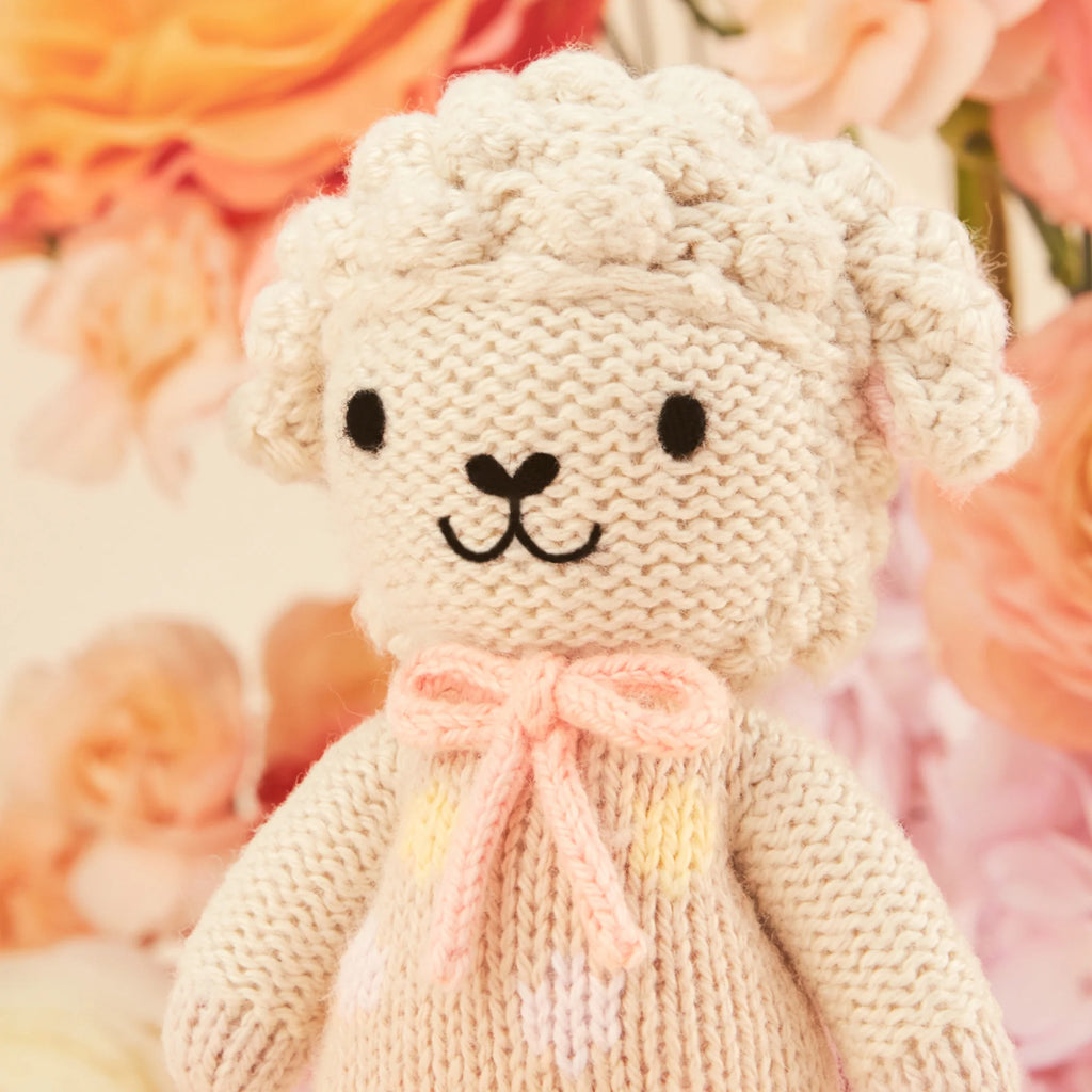 Cuddle + Kind Tiny Lucy the Lamb is a knitted plush sheep with a curly texture and smiling face, set against soft pastel flowers. Part of our sustainable toys collection, it sports a pink ribbon around its neck and wears a light-colored sweater.