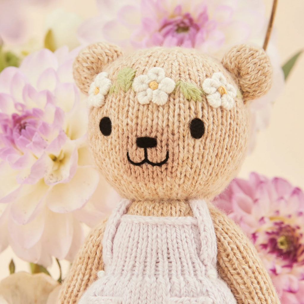 Tiny Olivia the Honey Bear by Cuddle + Kind, a hypoallergenic knitted teddy with a floral headband and white apron, stands smiling with black stitched eyes amid pink and white dahlias against a neutral background, offering gentle playtime fun.