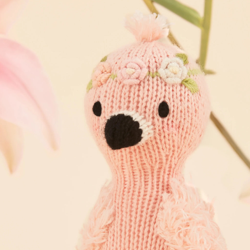 The Cuddle + Kind Tiny Penelope the Flamingo, knitted with floral embroidery on its head and a black beak, stands elegantly against a light background. Handmade in Peru, it showcases exquisite artistry with a soft pink flower partially peeking from the left side.