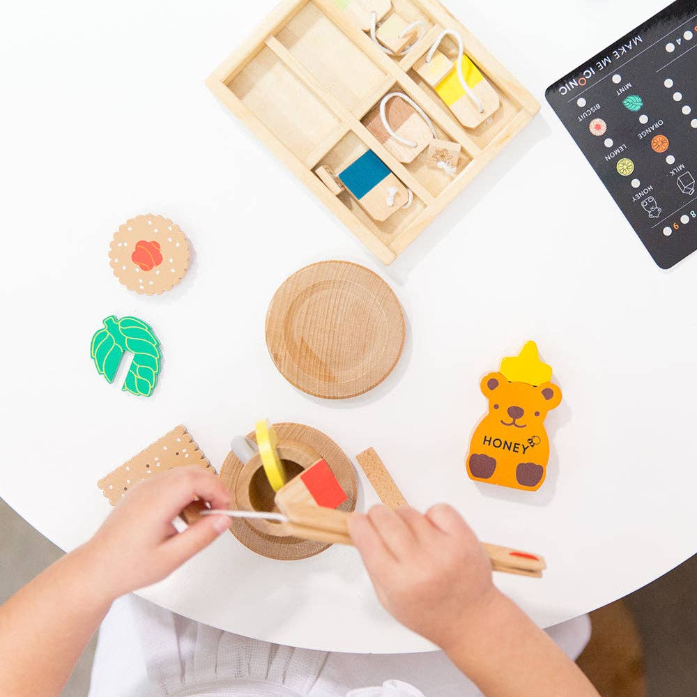 A child explores a play space with wooden food toys on a white table, featuring a honey bear, sandwich ingredients, and a small compartmentalized tray. Nearby, the Wooden Tea Set elegantly awaits an invitation to join an imaginary teatime adventure.