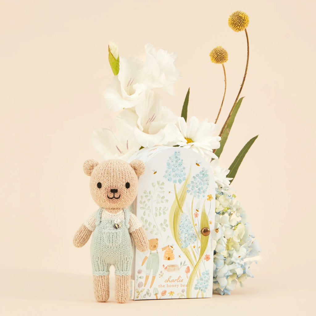 A Cuddle + Kind Tiny Charlie the Honey Bear, wearing mint green overalls and filled with hypoallergenic polyfill, sits next to a floral-themed book titled "Charlie the Honey Bear." Behind them is an arrangement of white flowers and hydrangeas against a light cream backdrop.