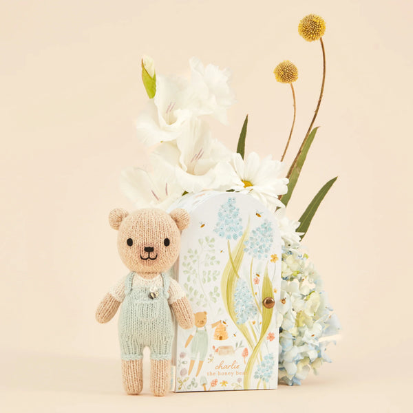 A Cuddle + Kind Tiny Charlie the Honey Bear, wearing mint green overalls and filled with hypoallergenic polyfill, sits next to a floral-themed book titled "Charlie the Honey Bear." Behind them is an arrangement of white flowers and hydrangeas against a light cream backdrop.
