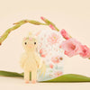 The handmade Cuddle + Kind Tiny Flora the Duckling (Mauve) from Peru stands before a decorative box with floral illustrations, framed by pink flowers and green leaves. This whimsical scene highlights sustainable fair trade practices.