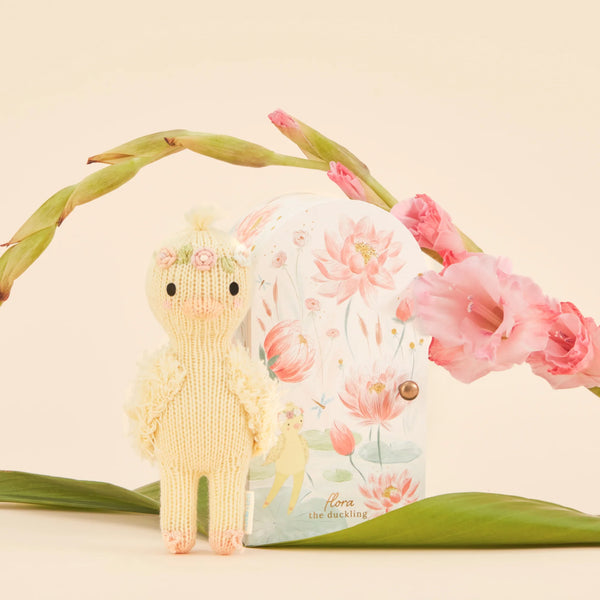 The handmade Cuddle + Kind Tiny Flora the Duckling (Mauve) from Peru stands before a decorative box with floral illustrations, framed by pink flowers and green leaves. This whimsical scene highlights sustainable fair trade practices.