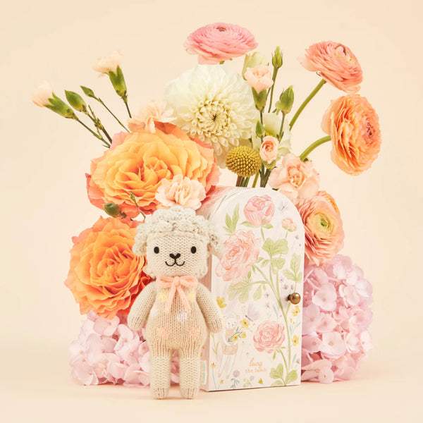The Cuddle + Kind Tiny Lucy the Lamb, a fair trade knitted toy, stands before a floral door-themed box amidst vibrant orange and pink flowers like roses and hydrangeas, against a light beige background.