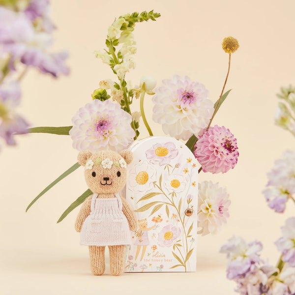 A Cuddle + Kind Tiny Olivia the Honey Bear, in a white dress and hypoallergenic design, sits next to a floral decorative box. In the background, a bouquet of pink and white flowers enhances the light beige setting.