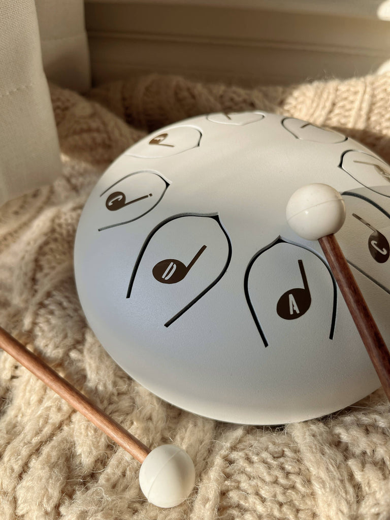 A white Steel Tongue Drum with labeled notes rests on a textured cream knit blanket, accompanied by two wooden mallets. Sunlight casts soft shadows across the relaxing musical scene.