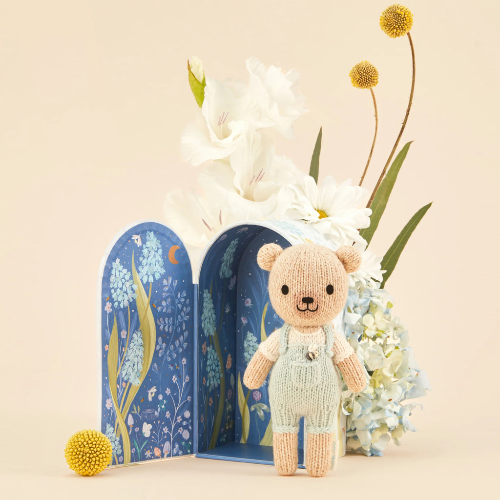 The whimsical scene of Cuddle + Kind's Tiny Charlie the Honey Bear in green overalls before a blue floral-patterned arch, adorned with white flowers and yellow billy buttons, is intricately crafted using fair trade practices, ensuring genuine care in every detail.