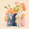 Cuddle + Kind Tiny Lucy the Lamb, a knitted toy with a pink bow tie, stands before an open floral-patterned decorative book. Part of our sustainable toys collection, it rests amid pink and peach blossom bouquets.