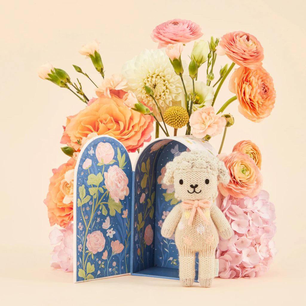 Cuddle + Kind Tiny Lucy the Lamb, a knitted toy with a pink bow tie, stands before an open floral-patterned decorative book. Part of our sustainable toys collection, it rests amid pink and peach blossom bouquets.