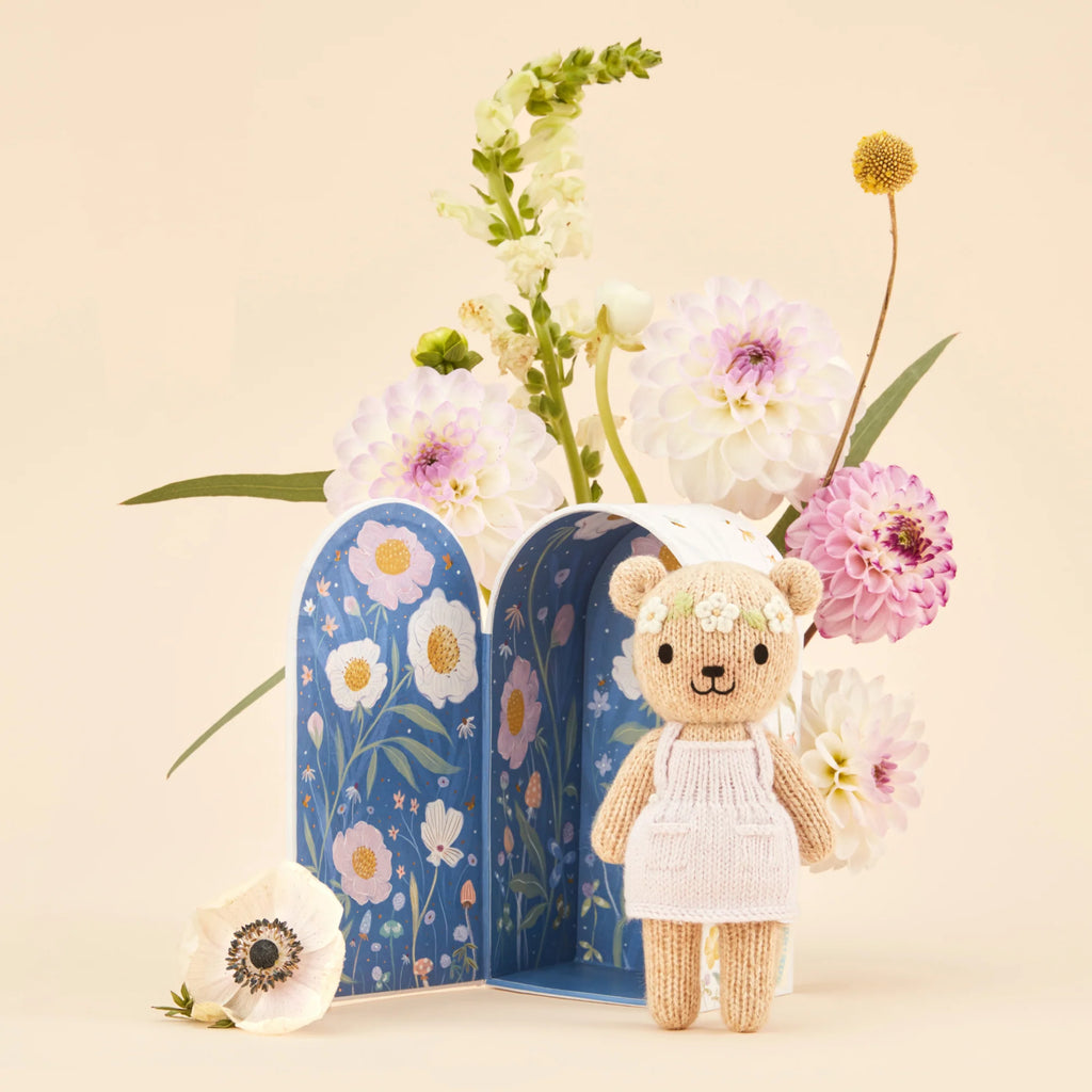 Tiny Olivia the Honey Bear, a hypoallergenic knitted teddy in a white dress, is posed before an open floral-patterned book with blooming flowers, including purple and white dahlias, against a light beige background.