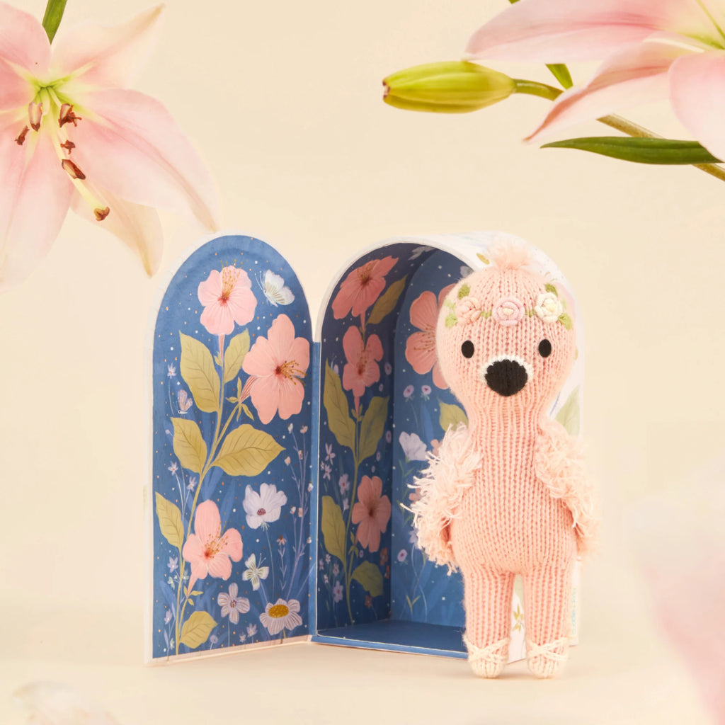 Cuddle + Kind's Tiny Penelope the Flamingo, a handmade pink stuffed animal from Peru, sports a floral headband. It stands in front of an open box with a blue background featuring pink and yellow floral designs, with pink flowers decorating the top corners of this charming scene.