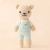 Cuddle + Kind Tiny Charlie the Honey Bear stands against a beige background, wearing light blue overalls and a white shirt. Stuffed with hypoallergenic polyfill, its black eyes and smiling mouth make it both adorable and safe for all.