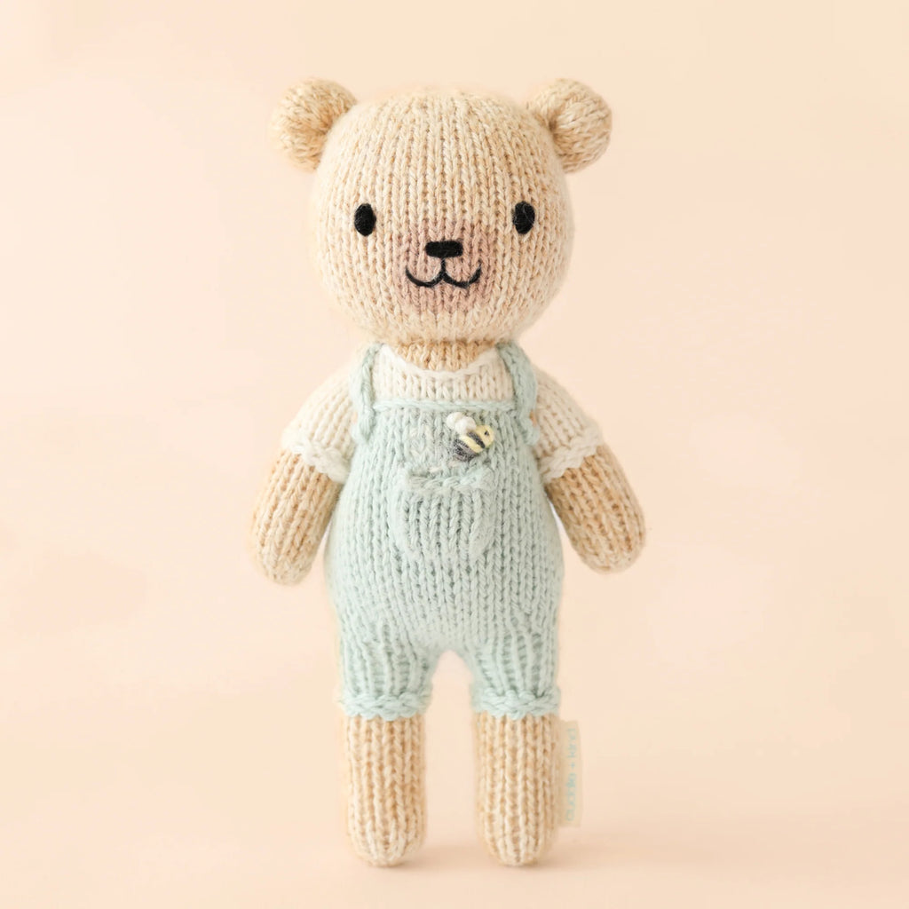 Cuddle + Kind Tiny Charlie the Honey Bear stands against a beige background, wearing light blue overalls and a white shirt. Stuffed with hypoallergenic polyfill, its black eyes and smiling mouth make it both adorable and safe for all.