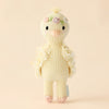 The Cuddle + Kind Tiny Flora the Duckling (Mauve) plush toy is handmade in Peru with fair trade practices. It features a fluffy knitted texture, small embroidered flowers on its head, and a cheerful expression with simple stitched eyes and beak against a soft, off-white backdrop.