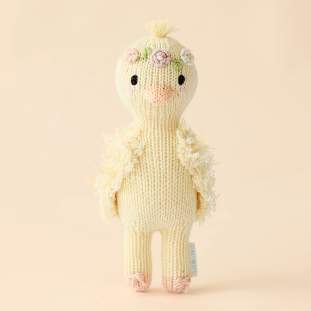 The Cuddle + Kind Tiny Flora the Duckling (Mauve) plush toy is handmade in Peru with fair trade practices. It features a fluffy knitted texture, small embroidered flowers on its head, and a cheerful expression with simple stitched eyes and beak against a soft, off-white backdrop.